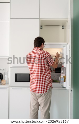 Delivery food, products to home. Shopping and healthy food concept. Young man in red plaid shirt holding disposable plastic boxed with food and putting them to the refridgerator