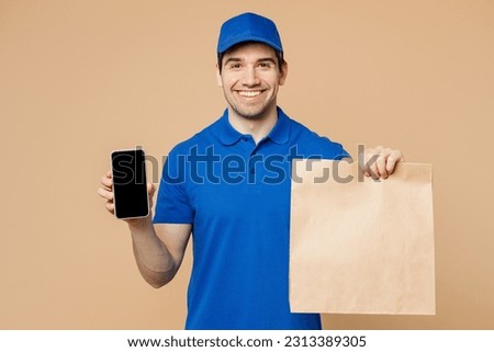 Delivery fun guy employee man wears blue cap t-shirt uniform workwear work as dealer courier hold brown clear takeaway bag use mobile cell phone blank screen area isolated on plain beige background