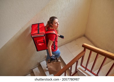 A delivery girl looks up as she goes up the stairs of a residential building carrying some bags of food and a backpack - Powered by Shutterstock