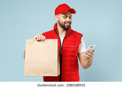 Delivery guy employee man in red cap white T-shirt vest uniform work as dealer courier service hold brown clear blank craft paper takeaway bag use cell phone isolated on pastel blue background studio