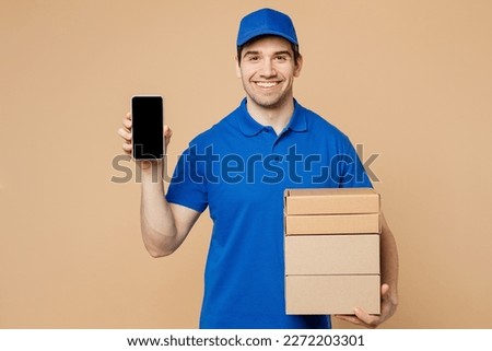 Delivery guy employee man wearing blue cap t-shirt uniform workwear work as dealer courier hold stack cardboard boxes use blank screen mobile phone isolated on plain beige background. Service concept