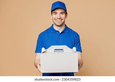 Delivery guy smiling fun employee man wears blue cap t-shirt uniform workwear work as dealer courier hold cake dessert in unmarked clear empty blank cardboard box isolated on plain beige background