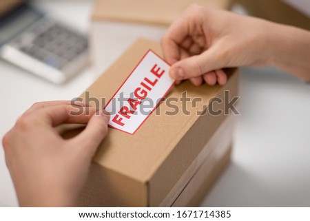 delivery, mail service, people and shipment concept - close up of woman sticking fragile mark to parcel box at post office