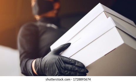 Delivery Man In Black Face Mask And Rubber Latex Nitrile Black Gloves Holding White Cardboard Box. Quarantine Concept.