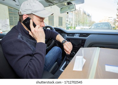 Delivery man in a cap is looking at his watch while talking on the phone in his van in front of the building. The package lies on the front seat.  - Powered by Shutterstock