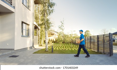 Delivery Man Holding Card Board Package Enters Through the Gates and Walks to the House. Delivering Postal Parcel. In the Background Beautiful Suburban Neighbourhood. Side View