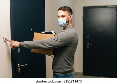 Delivery Man In Medical Mask Holding Cardboard Box And Contactless Payment POS Wireless Terminal For Card Paying, Ringing Doorbell Of Customer Apartment, Looking At Camera. Concept Of Online Shopping.