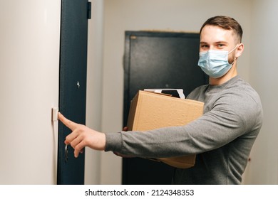 Delivery Man In Medical Mask Ringing Doorbell Of Customer Apartment Looking At Camera, Holding Cardboard Box And Contactless Payment POS Wireless Terminal For Card Paying, Concept Of Online Shopping.