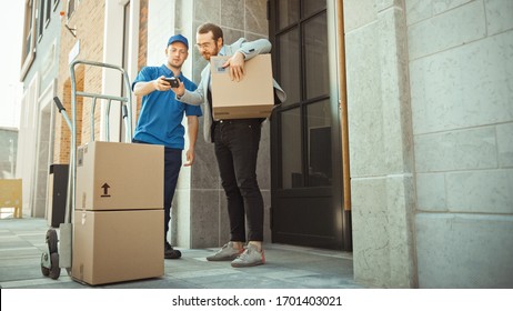 Delivery Man Pushes Hand Truck Trolley Full Of Cardboard Boxes Hands Package To A Customer, Who Then Signs Electronic POD Device. Courier Delivers Parcel To Man In Stylish Modern Urban Office Area