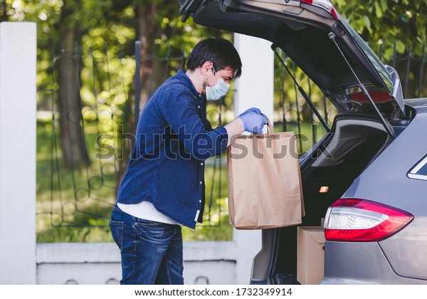 Delivery man putting paper bag\
with food in the car, food delivery man with protective mask and\
gloves