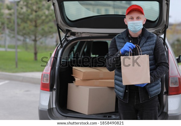 Delivery man in red cap, face medical mask and gloves\
hold paper bag near the car outdoor. Service coronavirus. Online\
shopping. mock up.