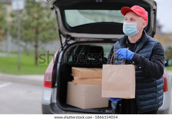 Delivery man in red cap, face medical mask and gloves\
hold paper bag near the car outdoor. Service coronavirus. Online\
shopping. mock up.