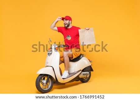 Delivery man in red cap t-shirt uniform driving moped motorbike scooter hold craft paper packet with food isolated on yellow background studio Guy employee working courier Service quarantine concept.