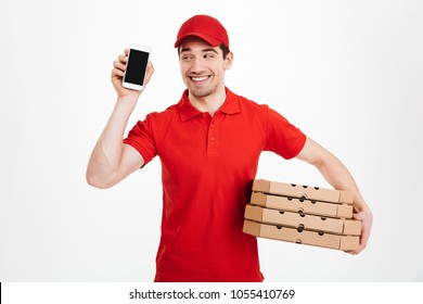 Delivery man in red t-shirt and cap holding stack of pizza boxes and showing copyspace screen of cell phone meaning call or text isolated over white background