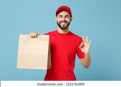 Delivery man in red uniform hold craft paper packet with food isolated on blue background, studio portrait. Male employee in cap t-shirt print working as courier. Service concept. Mock up copy space