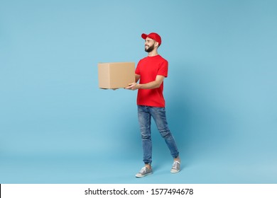 Delivery man in red uniform isolated on blue background, studio portrait. Male employee in cap t-shirt print working as courier dealer hold empty cardboard box. Service concept. Mock up copy space