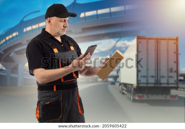 Delivery\
man with a tablet. Employee of a logistics company. Courier with\
box in his hands. Logistic man with a tablet. Delivery man near\
truck. Blurred road with truck in\
background.