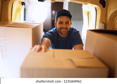 Delivery Man Takes Cardboard Package Out Of Truck. Delivery Guy Wearing Light Blue Uniform Reaching For Package To Take Out Of His Mini Van.