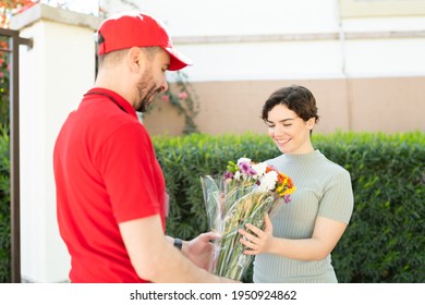 Delivery Man In A Uniform Delivering A Bouquet Of Flowers To A Beautiful Young Woman At Her Doorstep. Male Worker From A Flower Shop Making A Home Delivery