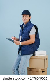 Delivery man in uniform is seating on a stack of parcel cardboard boxes over blue background. He is dressed in dark blue sleeveless jacket and cap. Filling up orders list.