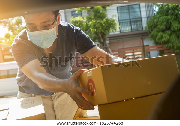 Delivery man wear mask looking box packages and\
picking up parcel from rear car trunk delivery, shipping, send to\
customer at home. New normal E-commerce shopping online form home\
delivery concept.