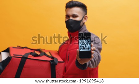 Delivery man wearing black mask holding red parcel box. Showing mobile phone screen in the camera with QR code. Concept of parcel delivery during Covid19 and digital pay. Yellow background studio shot
