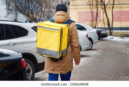 Delivery Man With Yellow Backpack Walking Down The City Street In Winter Season. Courier Of Delivery Food Service, Fast Deliver Groceries And Food Orders From Restaurants And Cafes. Takeaway Food