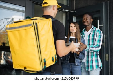 Delivery Man With Yellow Uniform And Cap That New Normal Style By Express Service, Holding And Sending Pizza Box Carton To Young Multiethnic Family Customers.