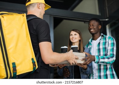 Delivery Man With Yellow Uniform And Cap That New Normal Style By Express Service, Holding And Sending Pizza Box Carton To Young Multiethnic Family Customers.