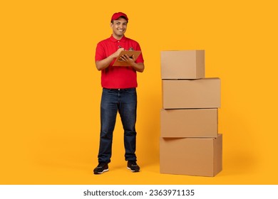 Delivery Offer. Hispanic Courier Man Holding Clipboard Filling Shipment Papers Near Carton Boxes Stack, Standing And Smiling To Camera, Posing With Packed Parcels On Yellow Background