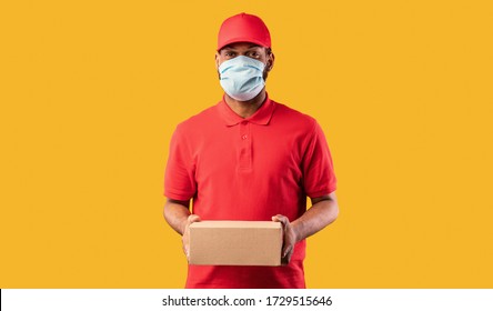 Delivery Service. African Courier Guy Holding Cardboard Box Delivering Parcel Package Wearing Protective Mask Posing Over Yellow Background. Studio Shot