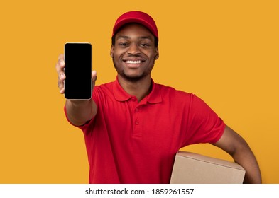 Delivery Service Application. Smiling African American Man Showing Smartphone With Blank Screen For Mockup, Wearing Red Cap, Uniform And Thermo Backpack Bag, Holding Box, Isolated On Orange Background