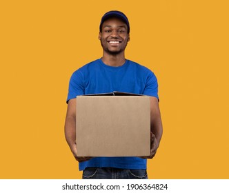 Delivery Service Concept. Smiling Black Mailman Wearing Blue Cap And T-shirt Holding Cardboard Box, Showing And Giving It To Camera, Copy Space. Postal Worker Standing On Orange Studio Background