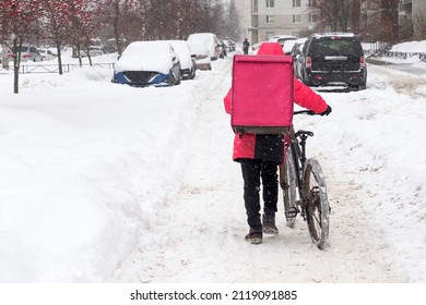 Delivery Service Courier With A Thermos Bag With Bicycle Walking Down The Snow In Winter City. Food Delivery Home