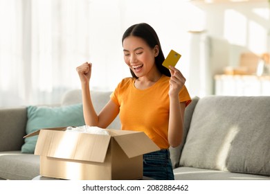 Delivery Service And Online Payment Concpet. Overjoyed Asian Lady Opening Box, Making YES Gesture And Holding Credit Card, Satisfied With Her Ordered Product, Sitting On Sofa