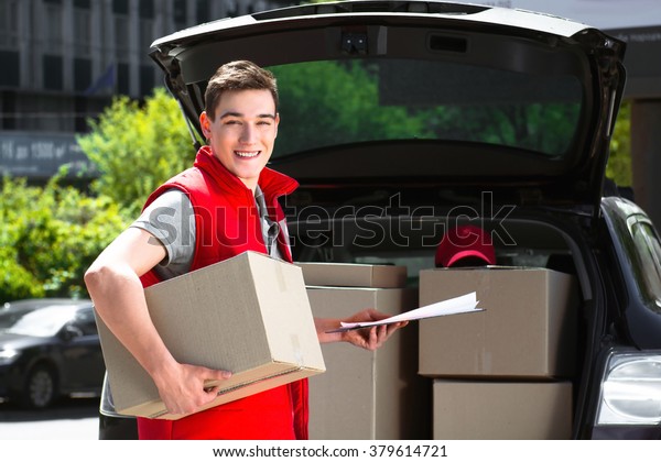 Delivery service worker in uniform delivering\
parcels and standing near car. Man holding box and document to\
sign, smiling and looking at\
camera