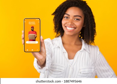 Delivery Tracking. Smiling African American Woman Holding And Showing Smartphone With Service App To Track Number, Courier Guy With Present Box On Screen, Isolated On Orange Background