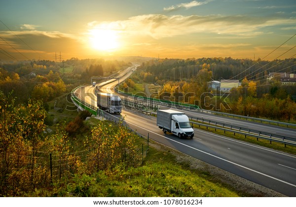 Delivery van and\
truck driving on the highway winding through forested landscape in\
autumn colors at\
sunset