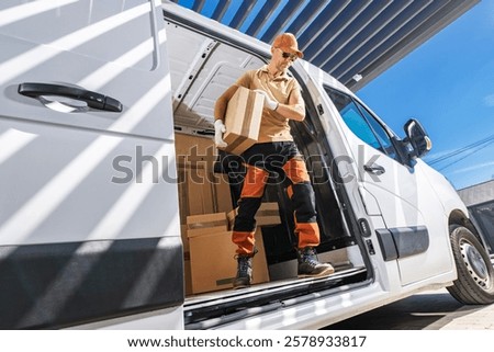 A delivery worker in a cap is unloading boxes from a van during the day, surrounded by clear skies and modern architecture.