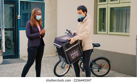 Delivery worker with face mask giving fastfood order to african american client, food delivery during coronavirus pandemic. Female courier on bicycle delivering restaurant meal in paperbag. - Powered by Shutterstock