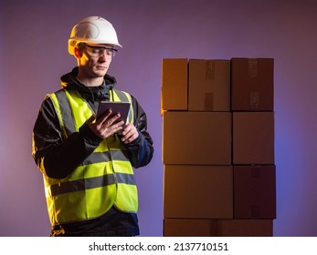 Delivery Worker. Man In Protective Helmet And Yellow Vest. Distributed Storage. Distribution Warehouse Worker. Delivery Service Employee On Dark Background. Several Cardboard Boxes Behind Him