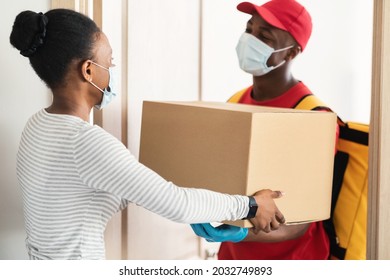 Deliveryman In Face Mask Giving Big Cardboard Box To Black Female Customer Standing Indoor. Courier Delivering Parcel To Lady's Home. Door Delivery Service Concept. Transportation And Shipping