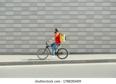 Deliveryman on bicycle. Young guy with beard and big backpack rides along path in city on gray brick wall background, side view, free space