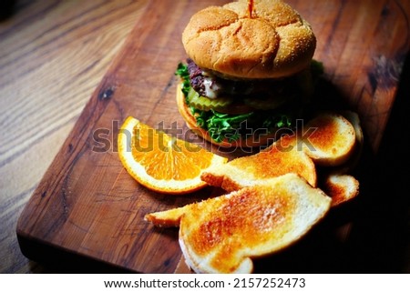 Deluxe Burgers, Hamburgers are being decorated before serving on a wooden cutting board consisting of toasted bread and orange slices.