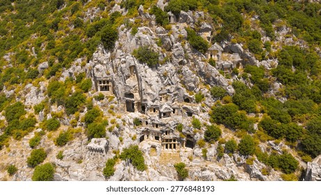 Demre, Turkey. The dramatic facade of the ancient Lycian rock-cut tombs at Myra, carved directly into a vertical limestone cliff face. Multiple tiers of temple-front and house-style tomb facades, complete with Ionic columns, carved doorways, and pediments, are stacked across the sunlit rock wall. The tombs replicate wooden Lycian domestic architecture in stone, preserving architectural forms that vanished elsewhere millennia ago. Dense green Mediterranean scrub and trees frame the grey and honey-toned cliff, creating strong color contrast. The scene is captured from an aerial drone perspective in bright summer daylight under clear skies, emphasizing the monumental scale and intricate stone carving of this ancient necropolis. The higher placement of the most elaborate tombs reflects the Lycian belief that elevation brought the deceased closer to the gods. This archaeological site, once part of the powerful League, remains one of the finest surviving concentrations of Lycian funerary architecture in Turkey.