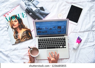 DENIA, SPAIN - OCTOBER 09, 2018. Young Girl Works On A Laptop On Bed. Magazines Vogue And Cosmetics On Background.