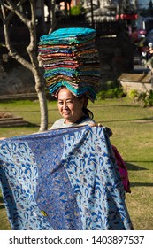 Denpasar, Bali / Indonesia - September 21 2017: Portrait Of A Smiling Middle Aged Balinese Woman. She Carries Huge Pile Of Colorful Sarongs On Her Head, Presents And Sells Them To Tourists.