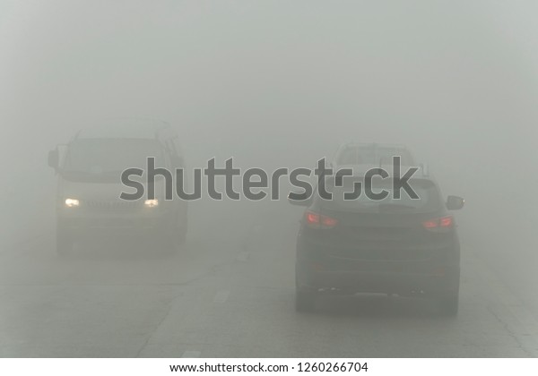 Dense fog and poor visibility on the road. Dangerous\
driving situations. View on highway traffic. Misty morning. Low\
visibility. Smoke on the road. cars driving trough mist. emergency\
danger on road
