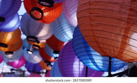 Dense overlapping lanterns viewed from below, with prominent orange and blue ones in the foreground and layers of purple, pink, and white behind, against a bright sky.