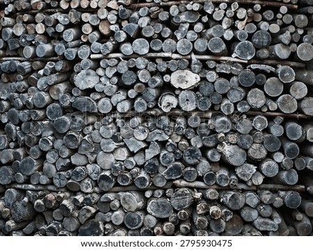 Dense stack of cut tree logs arranged horizontally in rows, showing circular end grain, bark texture, and varied diameters. The wood displays weathered gray